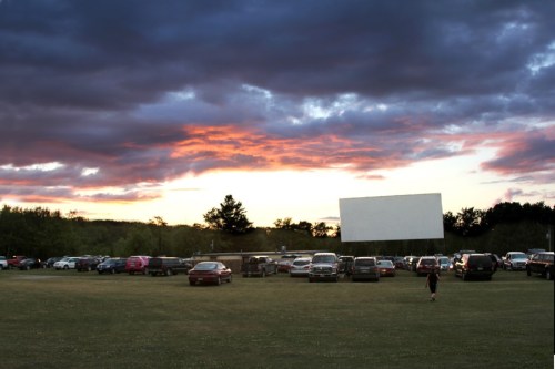 A drive-in movie theater at sunset, with cars parked on a grassy field and a large blank screen.