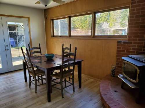 A cozy dining area with a wooden table, chairs, and a brick fireplace, featuring large windows and natural light.