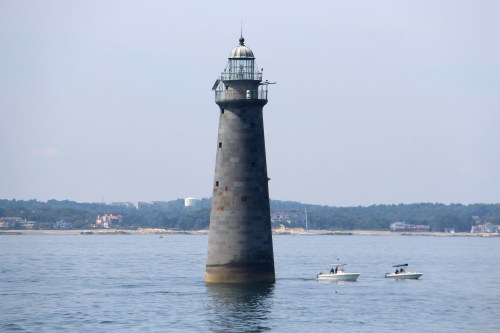 A tall, gray lighthouse stands in calm waters, with two small boats nearby and a distant shoreline in the background.