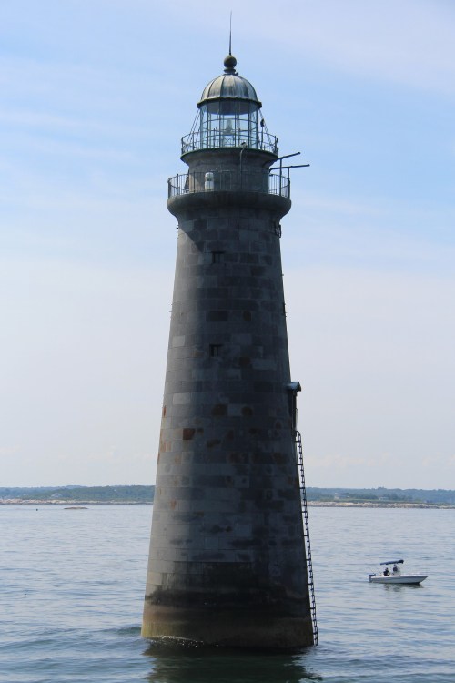 A tall, weathered lighthouse stands by the water, with a small boat nearby under a clear blue sky.