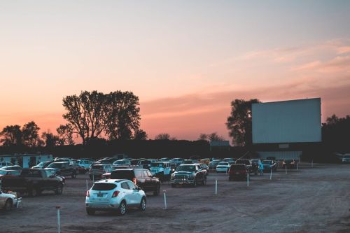 A drive-in movie theater at sunset, with cars parked in a gravel lot and a large screen in the background.