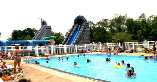 A busy pool area with people swimming and playing, featuring large inflatable water slides in the background.