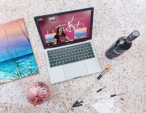 A laptop displaying a painting tutorial, surrounded by art supplies and a bottle of wine on a textured surface.