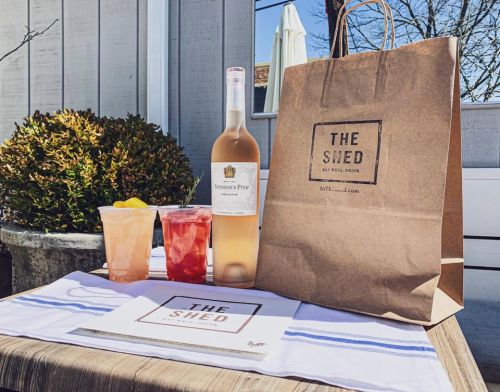 A bottle of rosé wine, two cocktails, and a takeout bag on a table with a menu, set outdoors.