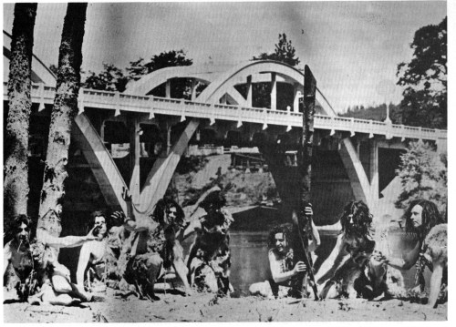 Historical black-and-white photo of people with dogs near a bridge, posing with sticks in a natural setting.