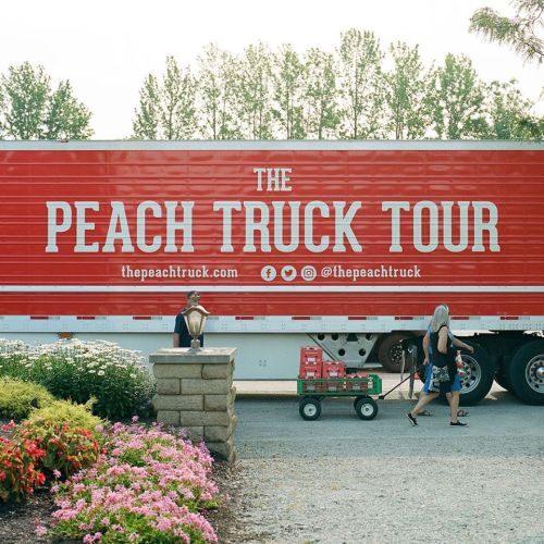 A red truck with "The Peach Truck Tour" logo parked near colorful flowers, with a person pulling a wagon nearby.