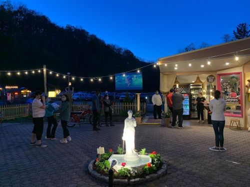 A lively outdoor scene at dusk with people gathered around a food stand and a fountain, string lights overhead.