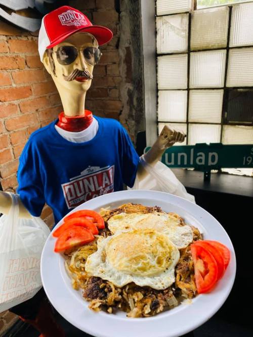 A mannequin in a blue shirt and red cap holds a plate of hash browns topped with two fried eggs and sliced tomatoes.