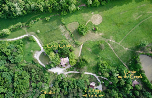 Aerial view of a lush green landscape featuring cabins, a studio, and a maze, surrounded by trees and a pond.