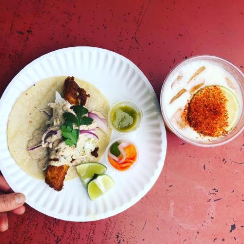A plate with a fish taco topped with onions and cilantro, accompanied by sauces, pickled vegetables, lime, and a drink.