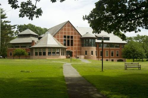 A large brick building with a unique architectural design, surrounded by green grass and trees, with a path and bench in view.
