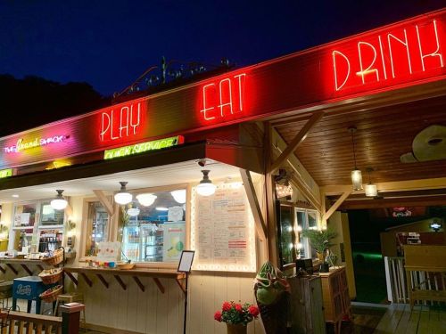 A brightly lit food shack at night, featuring neon signs that say "PLAY," "EAT," and "DRINK."