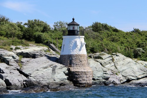 A white and stone lighthouse stands on rocky shore, surrounded by greenery and blue water under a clear sky.