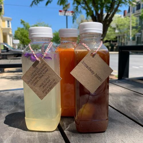 Three glass bottles of colorful drinks with tags, sitting on a wooden table outdoors. Bright sunlight and greenery in the background.