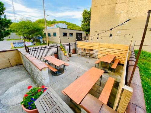 Outdoor seating area with wooden tables, string lights, and greenery, enclosed by a fence and adjacent to a building.