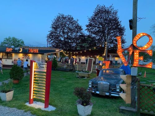 A lively outdoor scene at a drive-in with vintage cars, bright lights, and people enjoying the atmosphere at dusk.