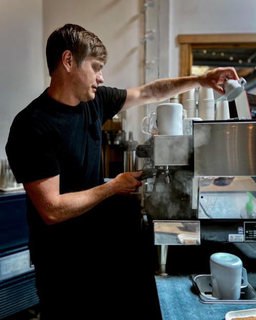 A barista prepares coffee at an espresso machine, pouring milk into a cup with steam rising around him.