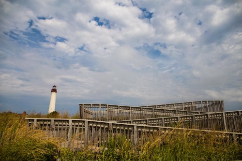 A wooden walkway leads to a lighthouse, surrounded by tall grass under a cloudy sky.