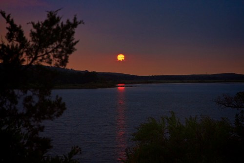 A serene sunset over a calm lake, with vibrant orange hues reflecting on the water's surface. Silhouetted trees frame the scene.