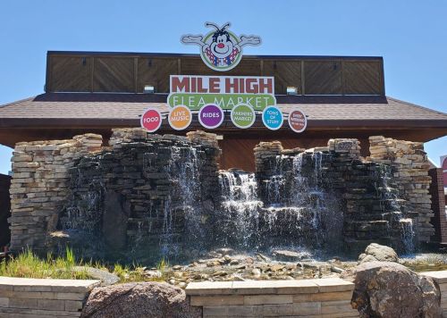 A colorful sign for Mile High Flea Market with a waterfall feature in front, promoting food, music, rides, and more.