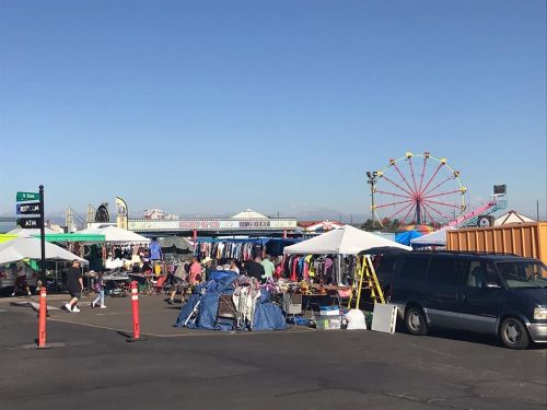 A bustling outdoor market with tents, clothing for sale, and a ferris wheel in the background under a clear blue sky.