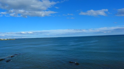 A serene ocean view under a bright blue sky with scattered clouds and gentle waves.