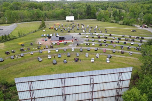 Aerial view of a drive-in theater with parked cars arranged in rows on a grassy field surrounded by trees.