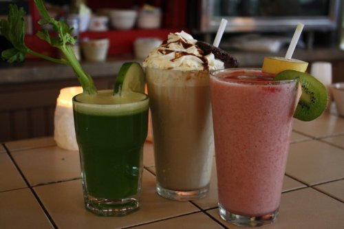 Three colorful drinks on a table: a green smoothie, a creamy dessert shake topped with whipped cream, and a pink fruit smoothie.