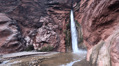 A waterfall cascades down a rocky cliff, surrounded by red rock formations and greenery, with a river at its base.