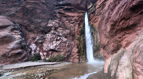 A waterfall cascades down a rocky cliff, surrounded by red rock formations and greenery, with a river at its base.
