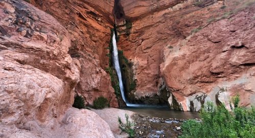 A waterfall cascades down red rock cliffs into a serene pool, surrounded by lush greenery and rugged terrain.