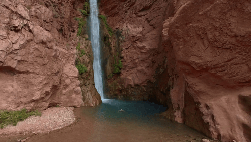 A serene waterfall cascades into a turquoise pool surrounded by red rock formations and lush greenery.