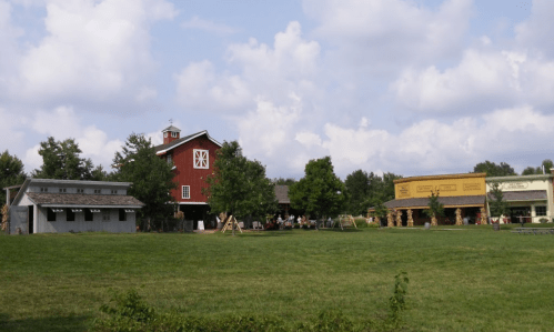 A red barn and nearby buildings surrounded by green grass and trees under a partly cloudy sky.