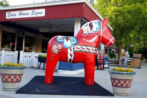 A large, colorful Dala horse statue stands outside a shop, surrounded by flower pots and greenery.