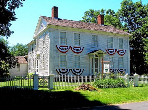 A historic white house adorned with red, white, and blue bunting, surrounded by greenery and a white picket fence.