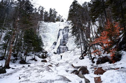 A person stands in front of a frozen waterfall surrounded by snow-covered rocks and trees, with autumn leaves nearby.