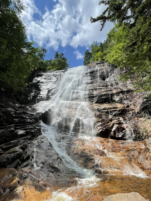 A cascading waterfall flows over rocky cliffs, surrounded by lush green trees and a bright blue sky with clouds.