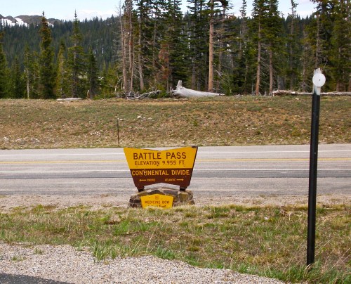 Sign at Battle Pass, elevation 9,955 ft, marking the Continental Divide with surrounding pine trees and mountains.