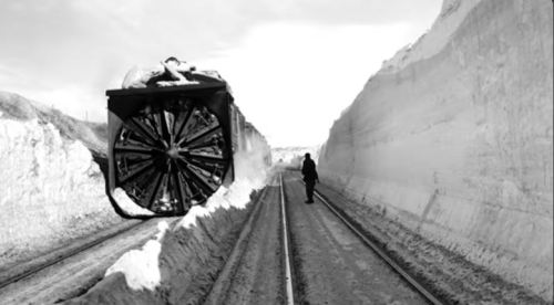 A snowplow train clears a path through deep snow, with a person standing nearby on the tracks. Black and white image.