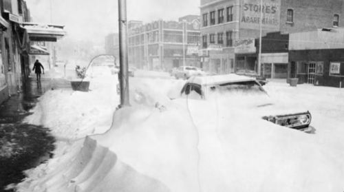 A snowy street scene with cars buried in snow and a person walking in a blizzard, creating a stark winter atmosphere.