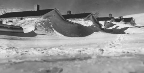 A snowy landscape with large snowdrifts surrounding a row of buildings under a cloudy sky.