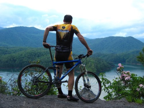 A cyclist in a yellow jersey stands on a trail, overlooking a scenic mountain lake surrounded by lush greenery.