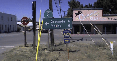 Road sign indicating directions to Grenada and Yreka, both 6 miles away, near a stop sign and a club.
