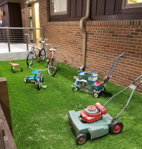 A small outdoor area with artificial grass featuring a bicycle, tricycles, and a lawn mower.