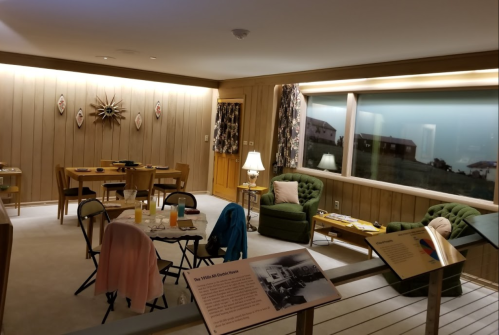 Mid-century modern living room with green chairs, wooden walls, and a dining area, showcasing retro decor and furnishings.