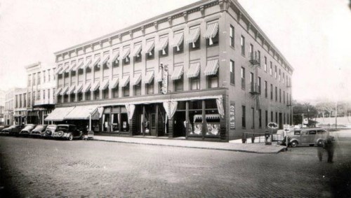 Historic building with awnings, vintage cars parked outside, and a street view from an earlier era.