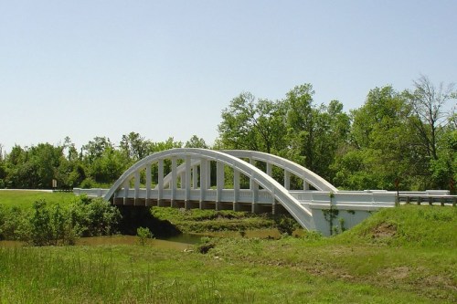 A white arch bridge spans a calm river, surrounded by green trees and clear blue skies.