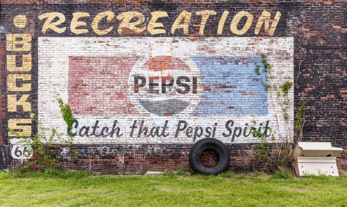 Faded Pepsi advertisement on a brick wall, with grass, a tire, and a white object in front.