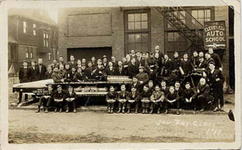A large group of men poses in front of a building, with a vintage car and "Cleveland Auto School" sign in the background.