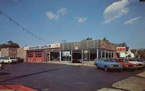 A vintage car dealership with a brick facade, featuring several parked cars and a clear blue sky.